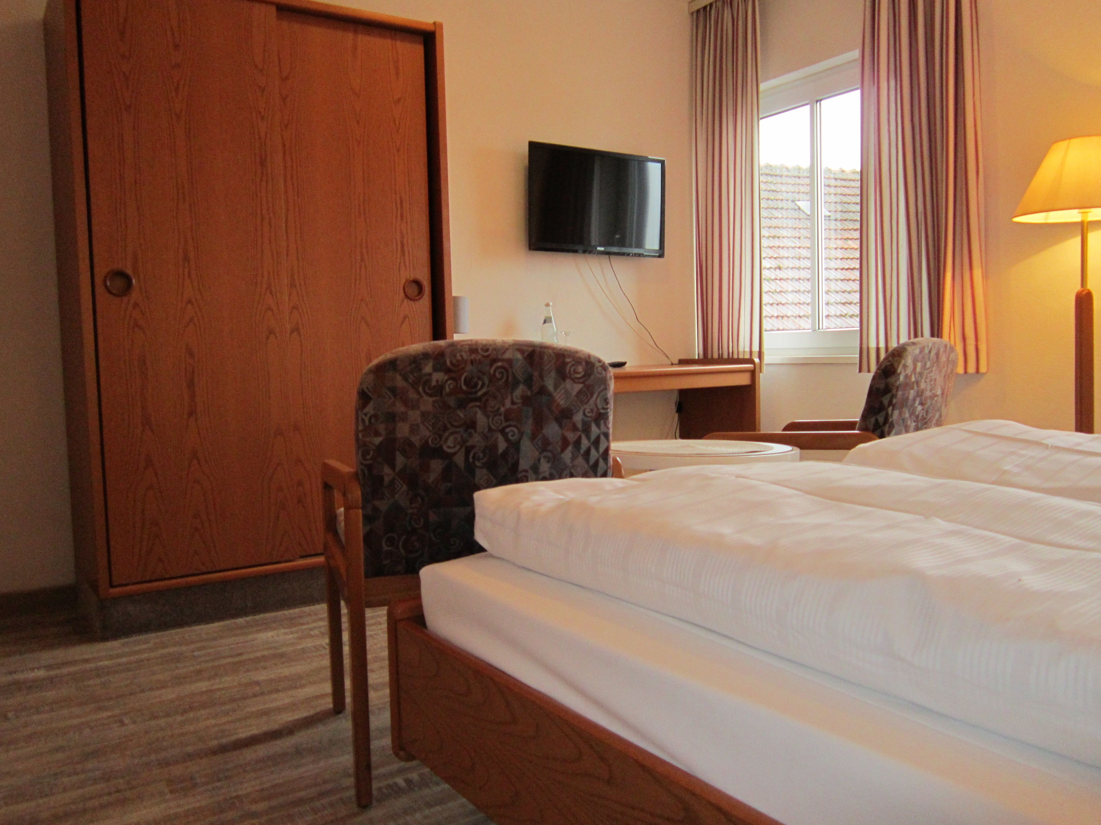 Une chambre d’hôtel avec une armoire en bois, un téléviseur fixé au mur, un bureau avec une chaise et un lit avec deux oreillers.