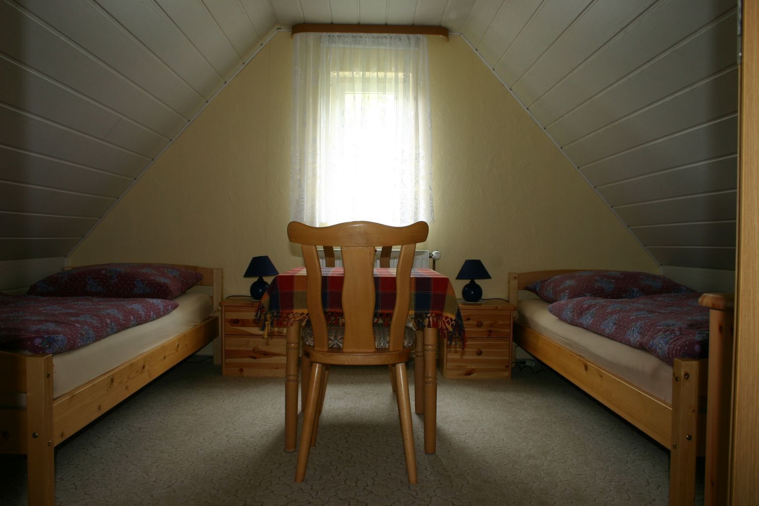 A small attic bedroom with two single beds, a wooden chair, a small table, and a window with sheer curtains.