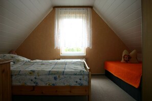 A bedroom with a sloped ceiling, a bed with floral bedding, a window with sheer curtains, and a small bedside table.