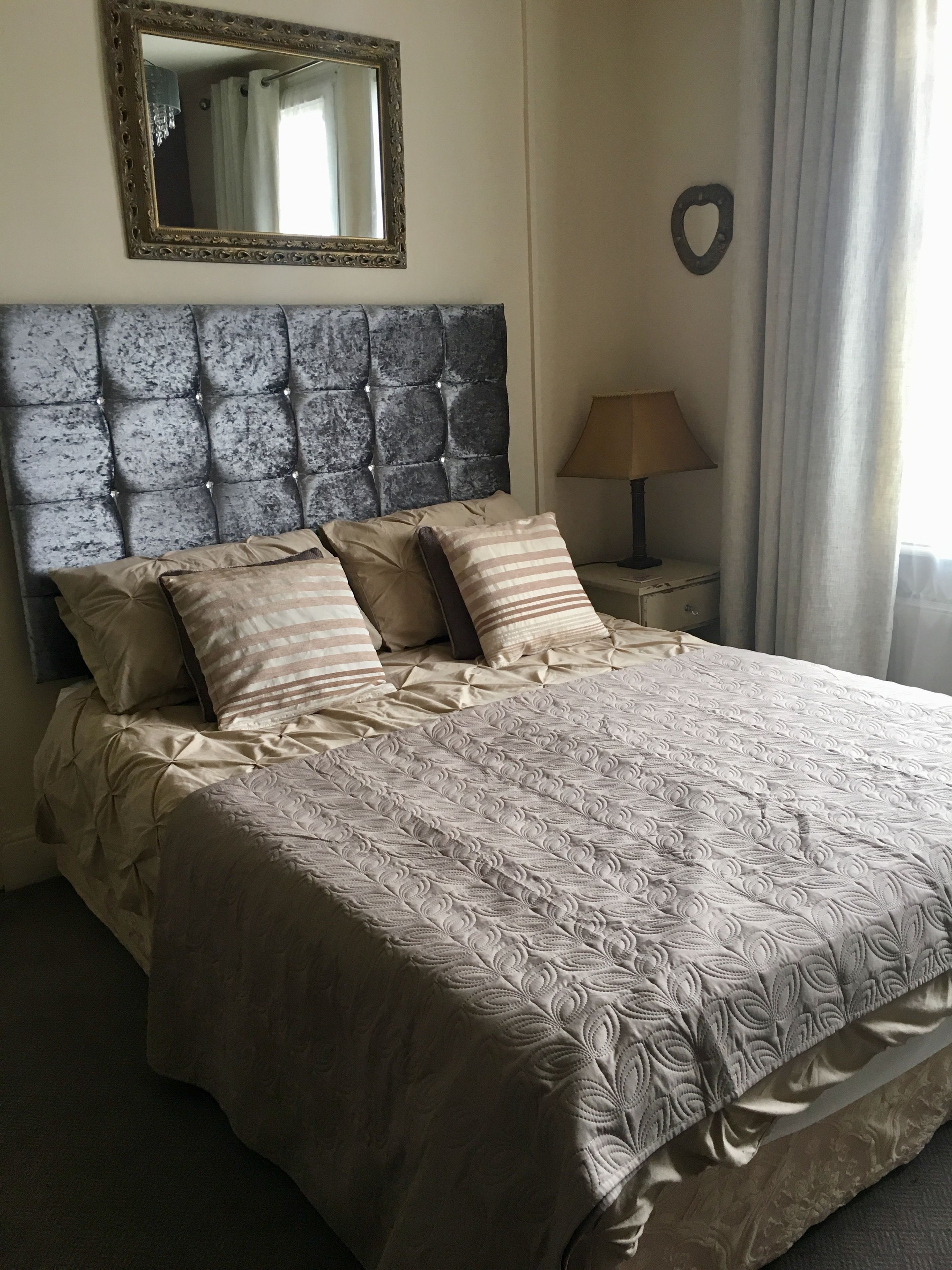 Una cama bien tendida con un cabecero capitonado, una lámpara de noche y un espejo en la pared.