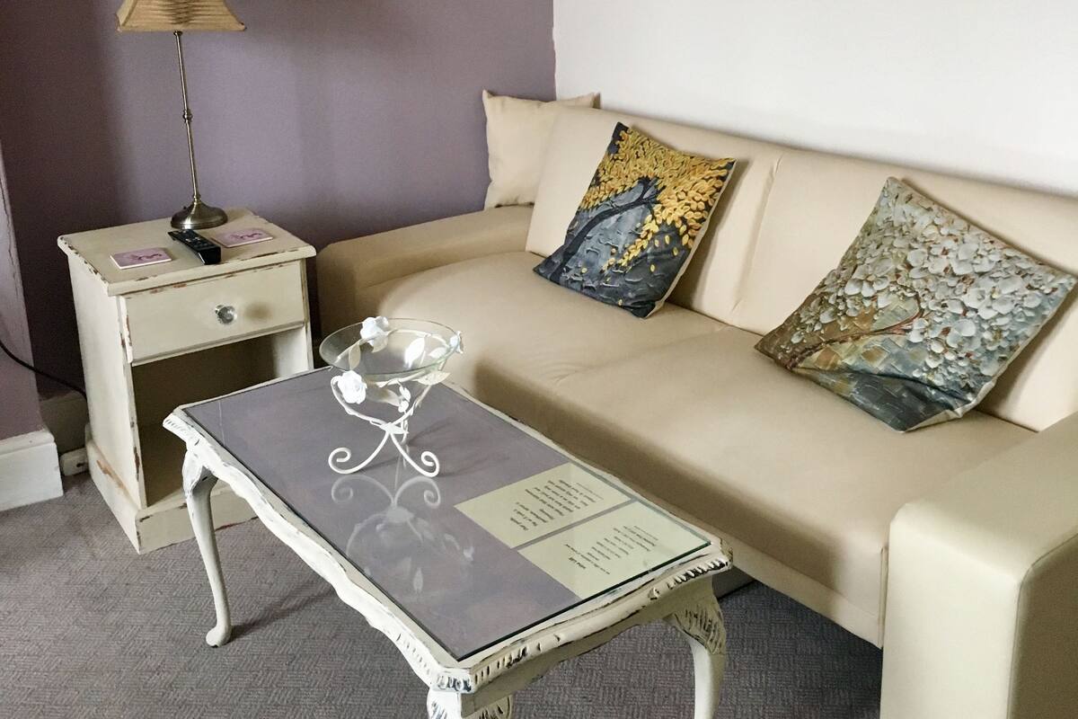 A room with a beige sofa, a small table with a glass top, a nightstand with a lamp, and a painting on the wall.