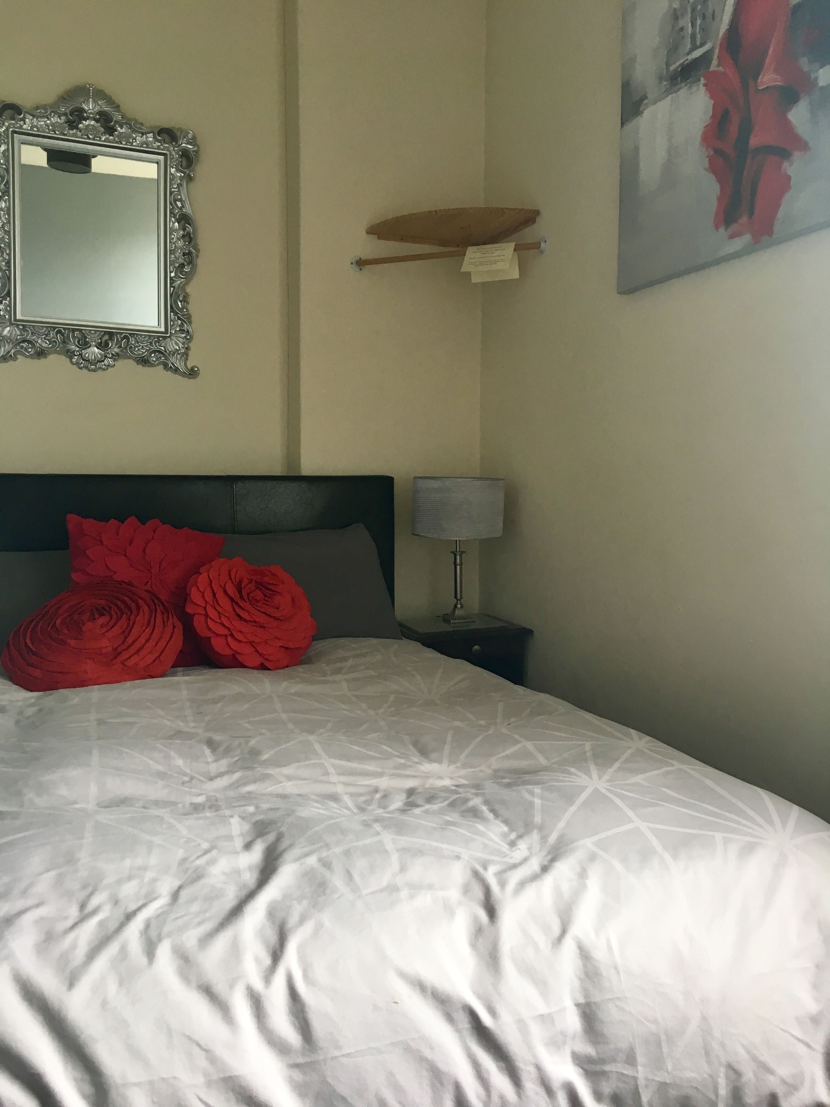 A neatly made bed with a white comforter and red rose-shaped pillows, a bedside lamp, a wall-mounted mirror, and a piece of artwork.