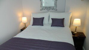A neatly made bed with a purple bedspread and pillows, flanked by two bedside tables with lamps, in a room with a decorative mirror above the bed. - Stoneway Guest House (Bridgnorth)