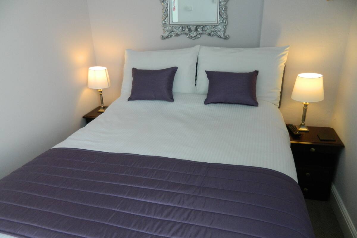 A neatly made bed with a purple bedspread and pillows, flanked by two bedside tables with lamps, in a room with a decorative mirror above the bed.