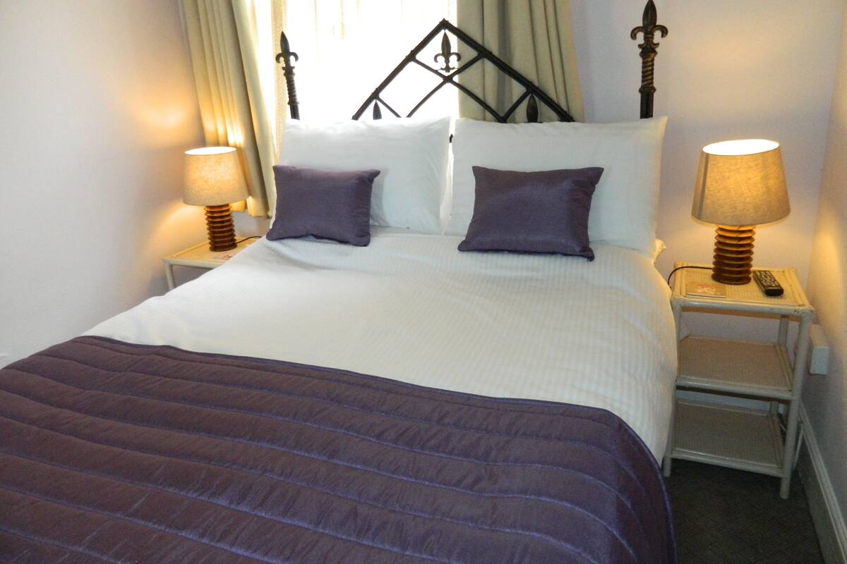 A neatly made bed with a dark purple bedspread and two pillows, flanked by two bedside tables with lamps and a remote control.
