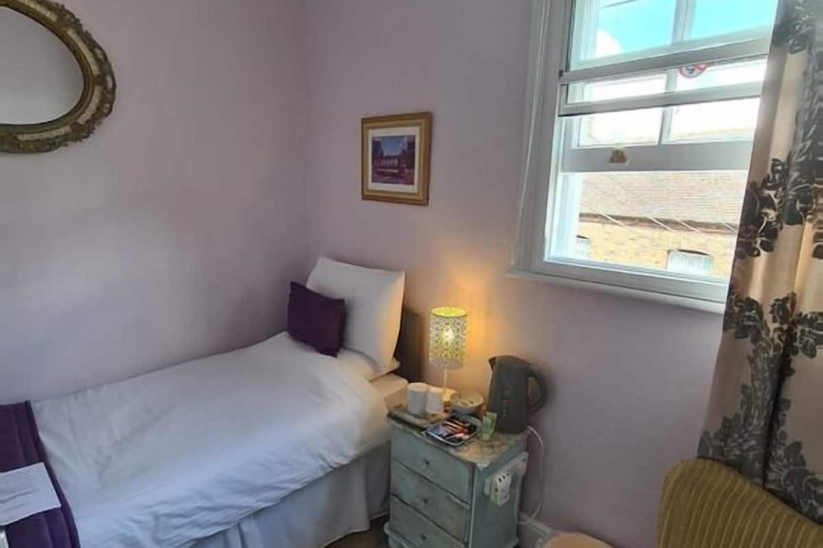 A bedroom with a bed, a nightstand, a lamp, a picture on the wall, and a window with a view of the outside.