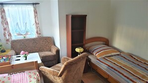 Um quarto pequeno e acolhedor, com uma cama de solteiro, uma poltrona, uma escrivaninha e uma estante de livros.