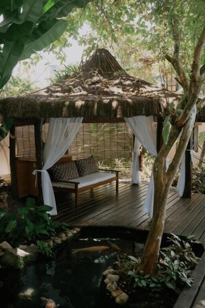 A wooden deck with a thatched gazebo, a bench, and a small pond.