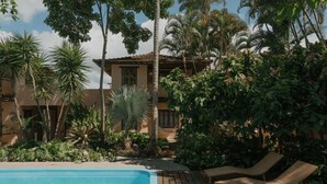 Un espace piscine agrémenté de chaises longues, entouré d’une végétation tropicale et bordé d’un bâtiment dont le toit est recouvert de tuiles.