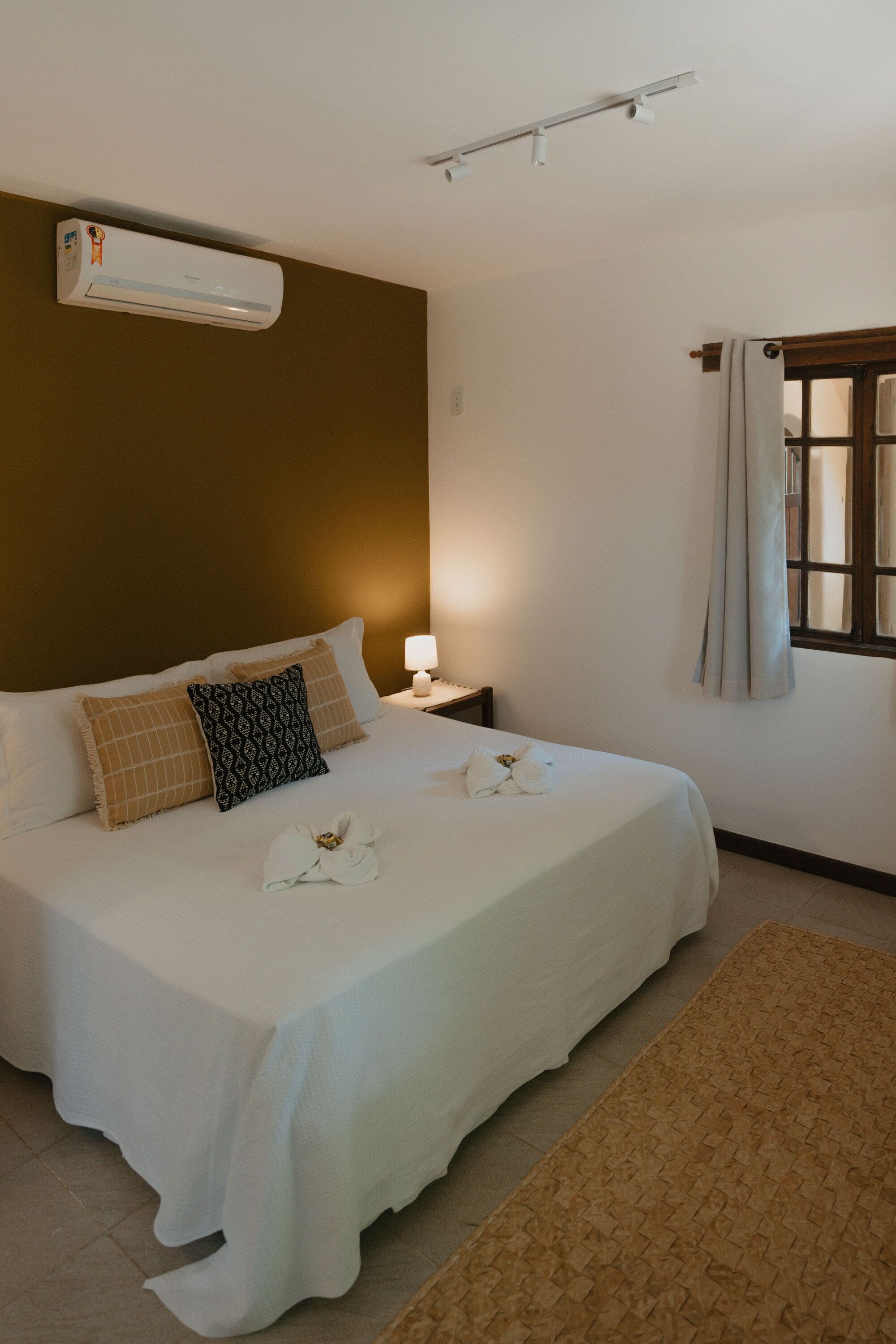 Una cama bien hecha con ropa blanca, pared marrón, ventana con cortinas y aire acondicionado instalado en la pared.
