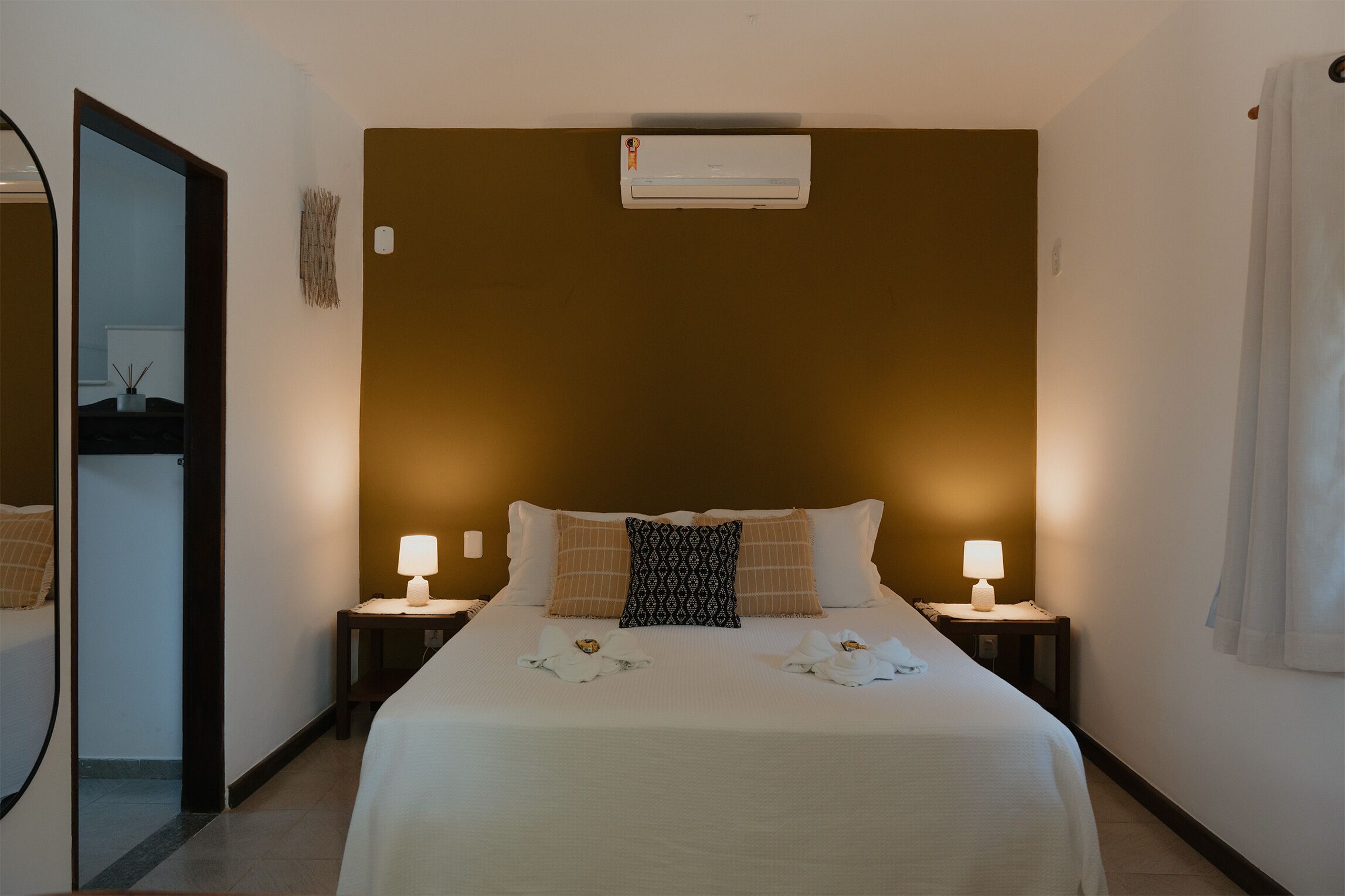 A hotel room with a bed, two bedside lamps, an air conditioning unit, a mirror, and a door leading to another room.