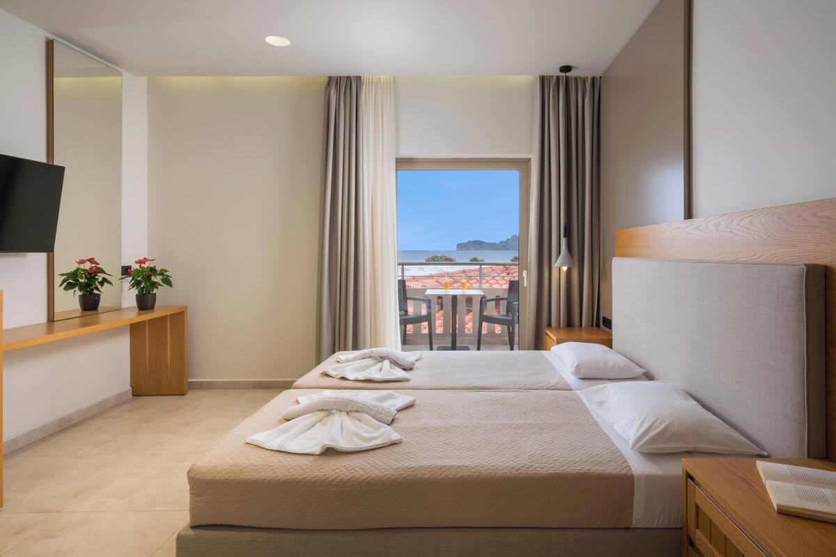 A hotel room with two beds, a TV, a desk with a computer, and a balcony with a table and chairs.