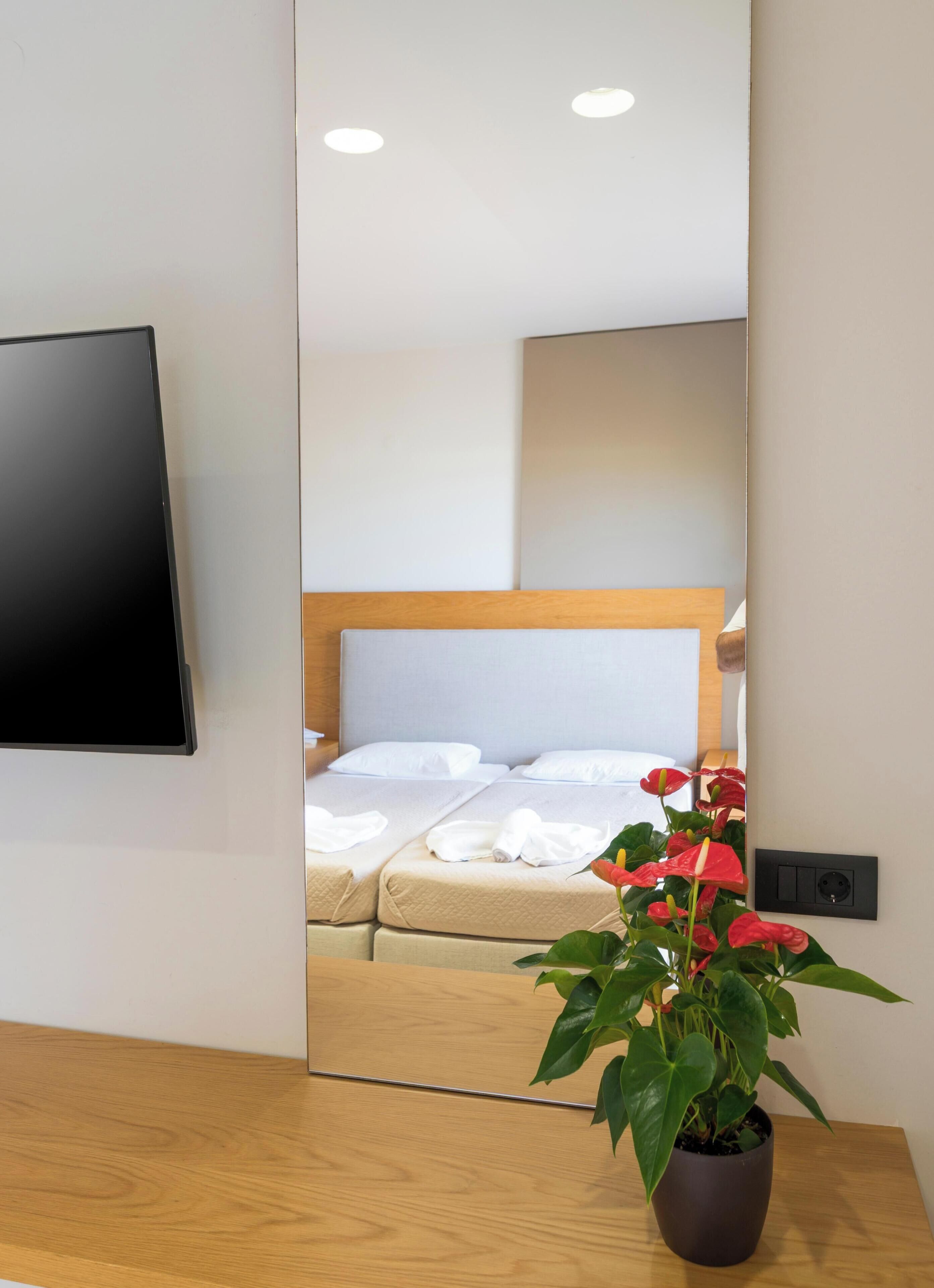 Une chambre d’hôtel dotée d’un grand miroir dans lequel se reflètent un lit, une tête de lit en bois et une plante en pot ornée de fleurs rouges.