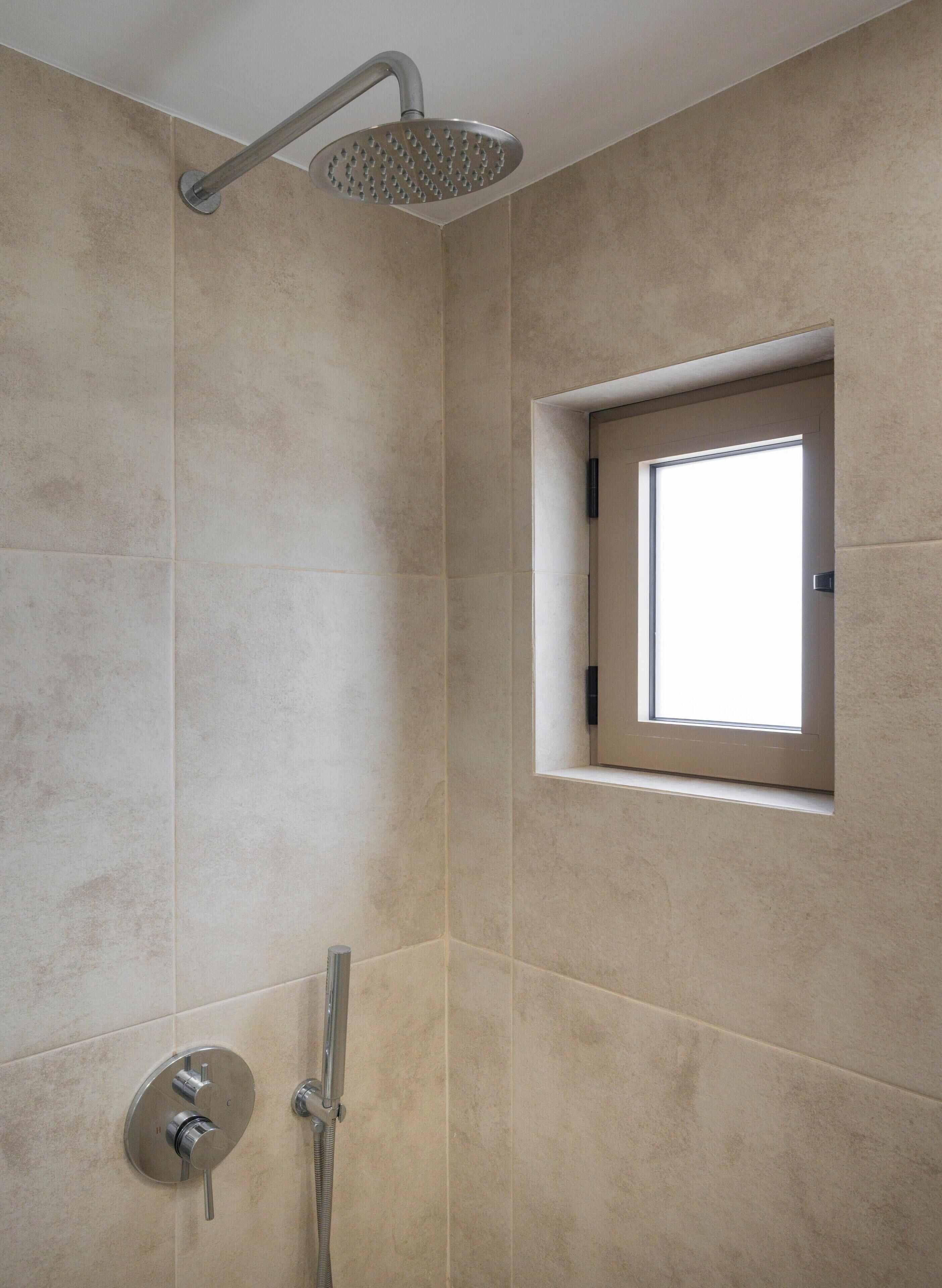 A shower with a rain showerhead and a handheld showerhead, a small window, and a wall-mounted control panel.