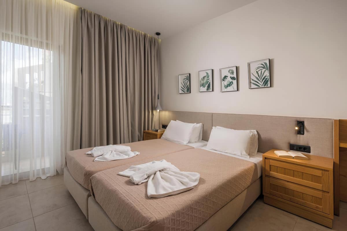 A hotel room with two beds, a large window with sheer curtains, a bedside table with a lamp, and four framed botanical prints on the wall.