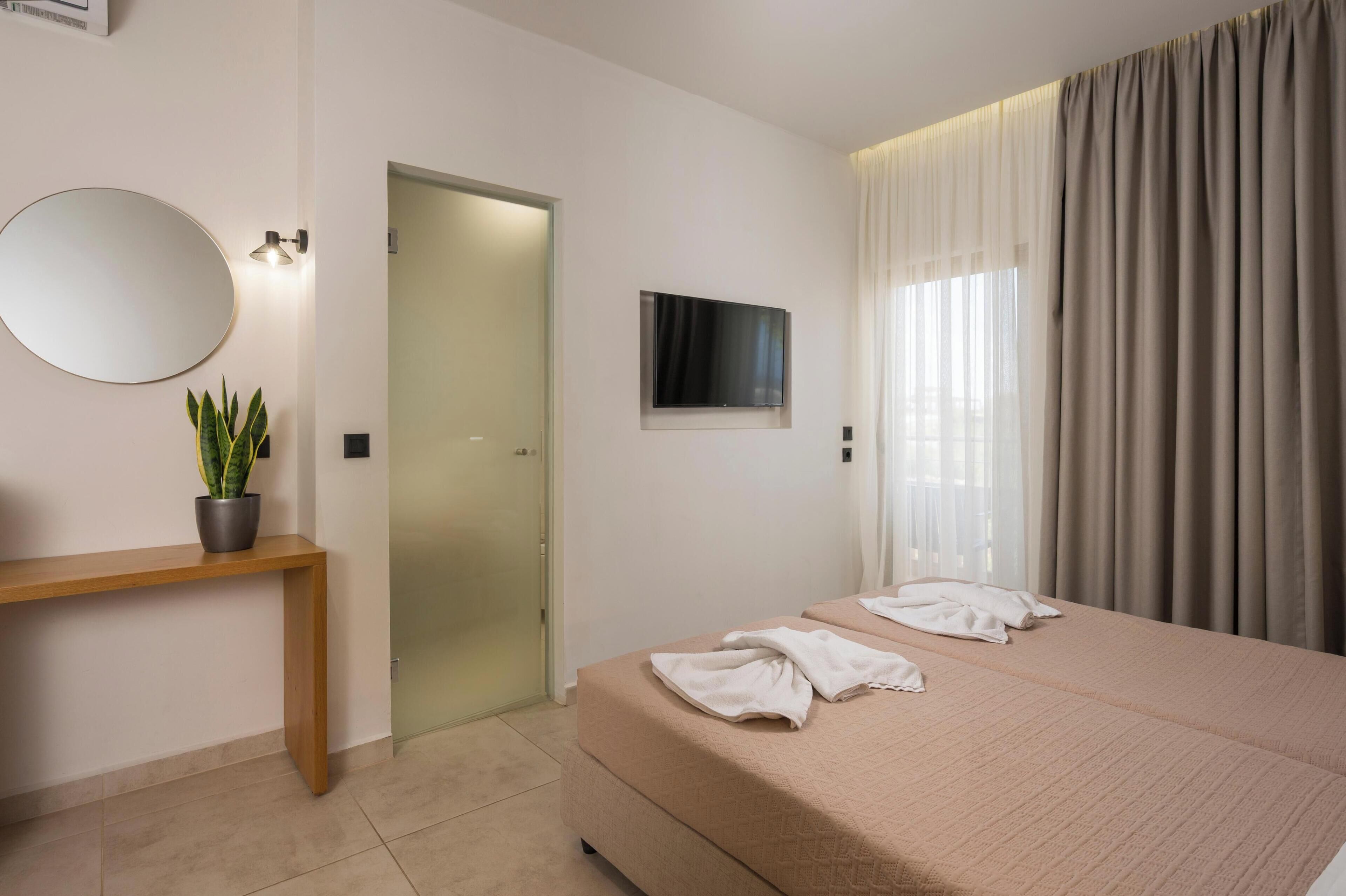 A hotel room with a bed, a TV, a mirror, a plant, and a wooden bench.