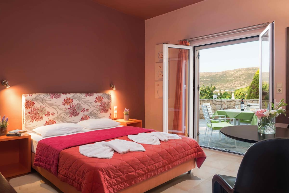 A hotel room with a bed, a nightstand, a table with a vase, and a view of the outdoors.
