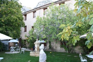 Une cour avec des tables et des chaises en pierre, une fontaine et un bâtiment doté d’un toit en tuiles.
