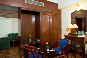 Une chambre d’hôtel avec une armoire en bois, un bureau avec une lampe, deux chaises, une petite table avec un vase et un canapé vert.