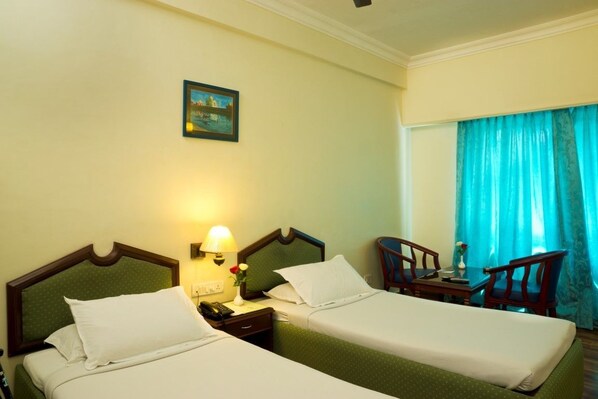 Habitación de hotel con dos camas individuales, una mesita de noche con lámpara y teléfono, una pequeña mesa con un jarrón y una ventana con cortinas.