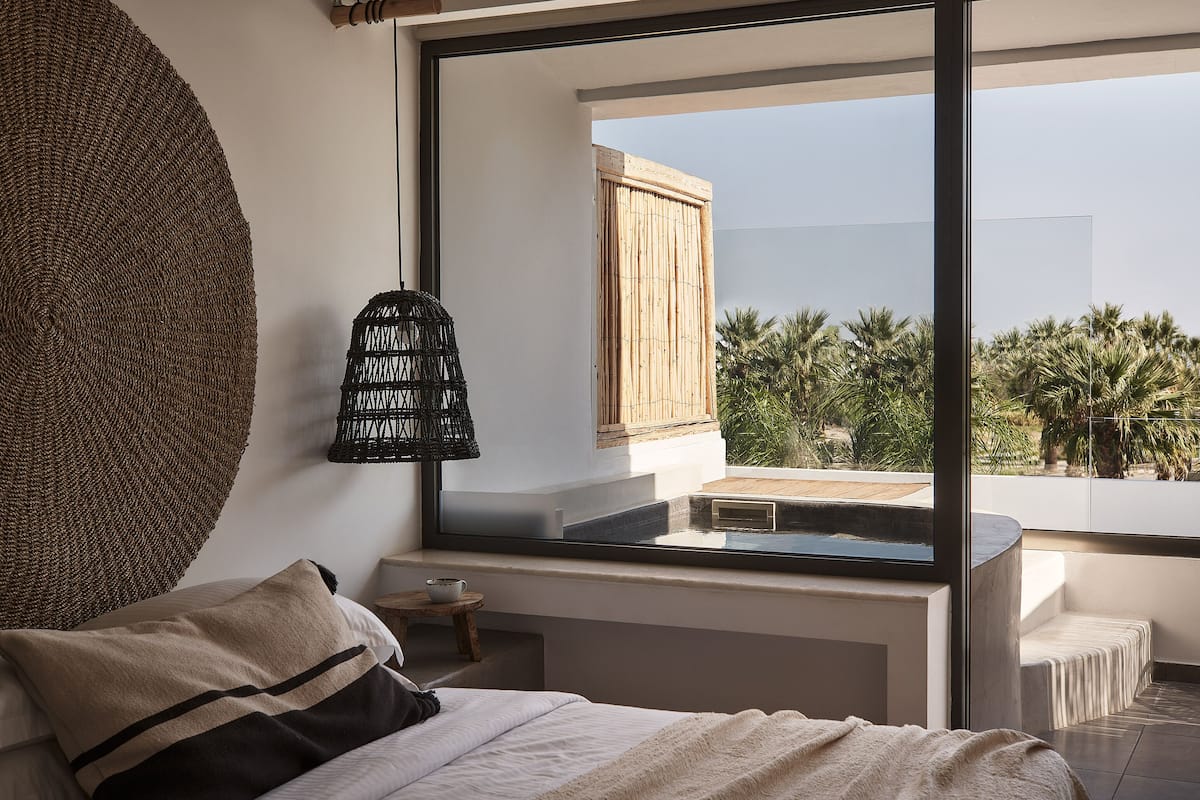 A bedroom with a large window, a bed, a small table, and a woven wall hanging.