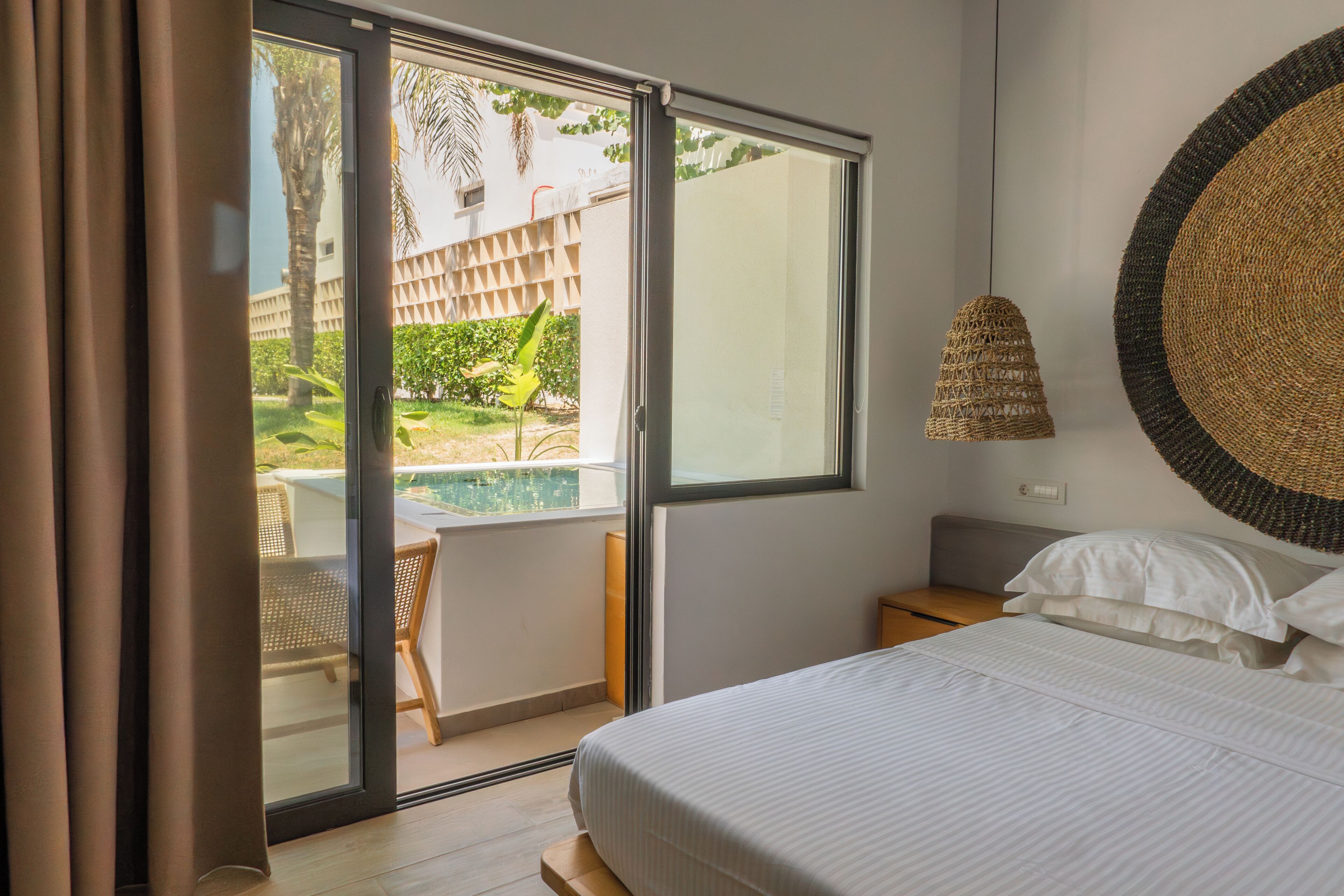 Habitación de hotel con cama, mesita de noche pequeña, lámpara colgante y puerta corrediza de vidrio que da a una zona de piscina.