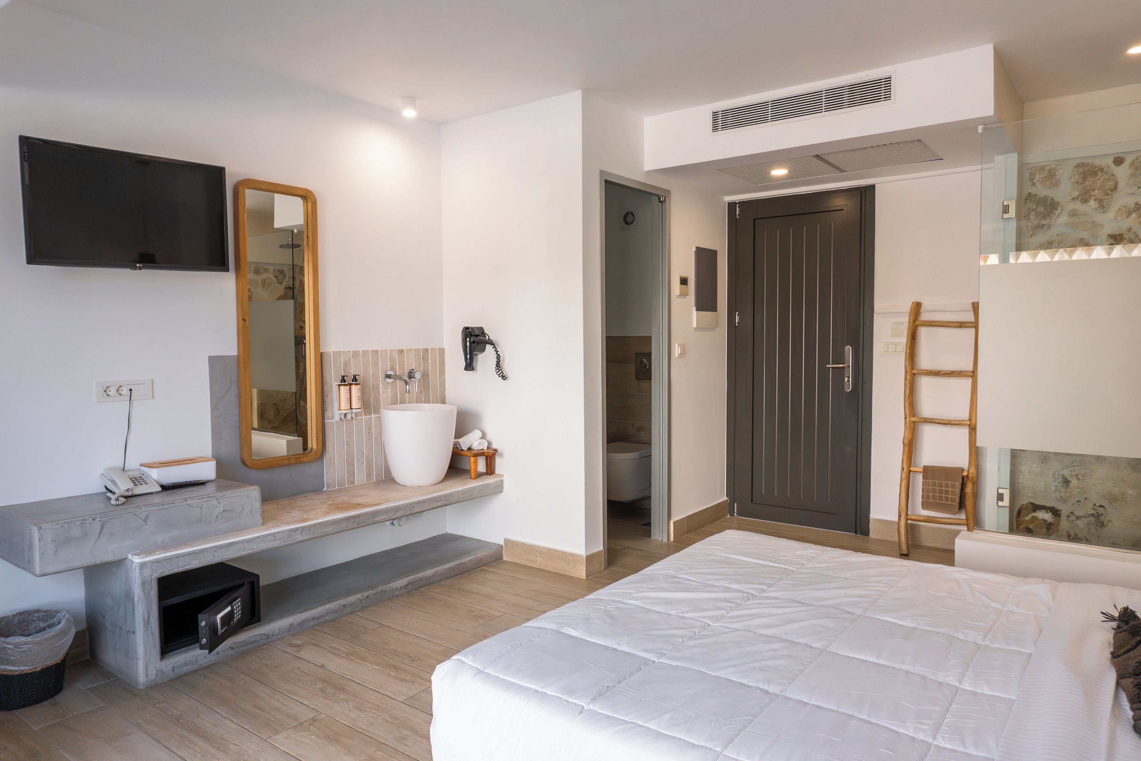 A modern bedroom with a large bed, a built-in bench with a mirror, a TV mounted on the wall, and a bathroom visible through an open door.