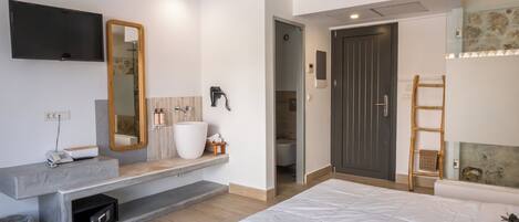 Une chambre moderne avec un grand lit, un banc intégré doté d’un miroir, un téléviseur fixé au mur et une salle de bain visible à travers une porte ouverte.