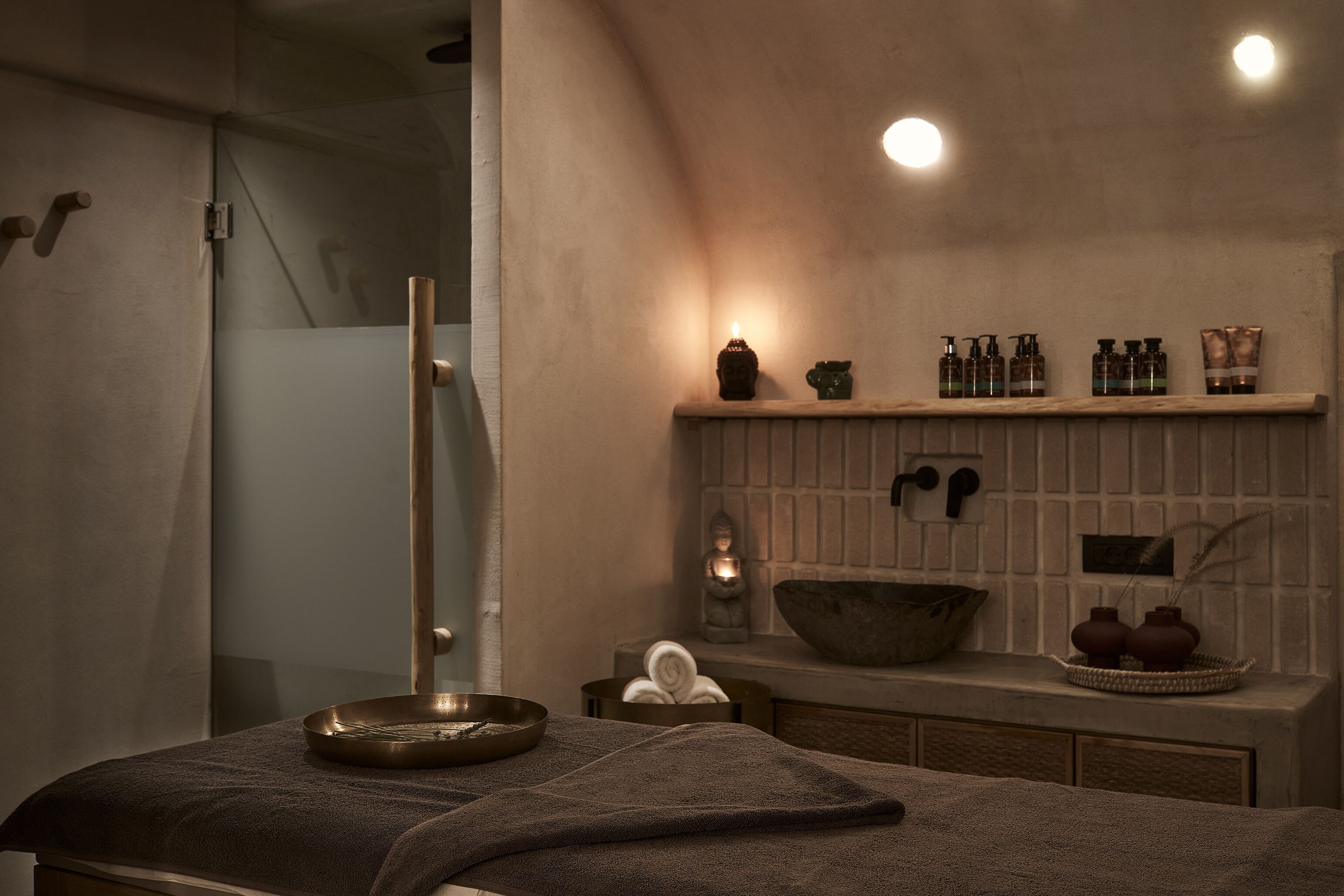 A modern bathroom with a glass shower enclosure, a wooden towel rack, a shelf with toiletries, and a stone basin.