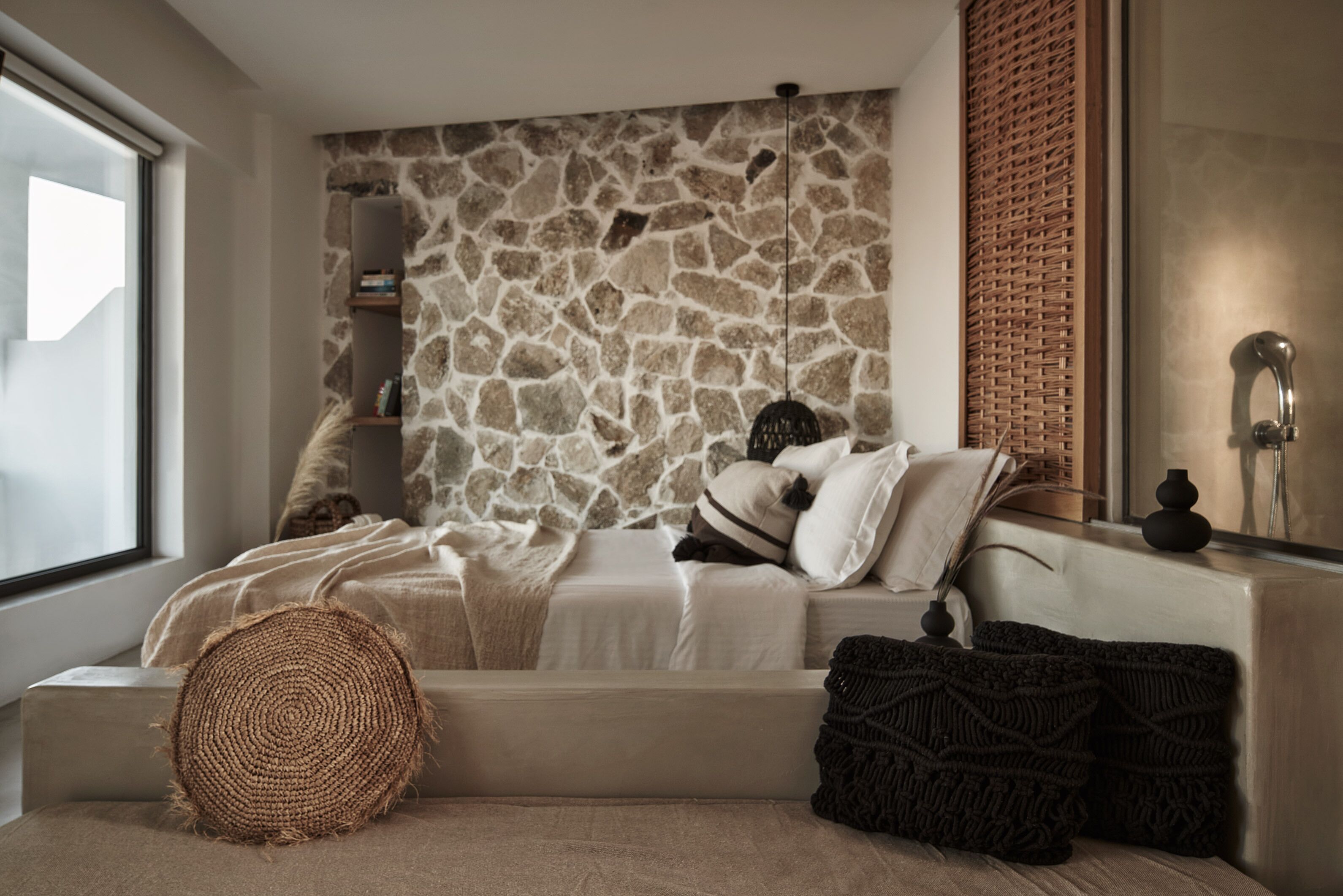Un dormitorio moderno con pared de piedra, un ventanal grande, una cama con varias almohadas y una mesita de noche con un jarrón.