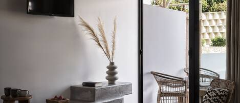 A modern bedroom with a flat-screen TV mounted on the wall, a concrete bench with decorative items, a wicker chair with a patterned pillow, and a sliding glass door leading to an outdoor area.