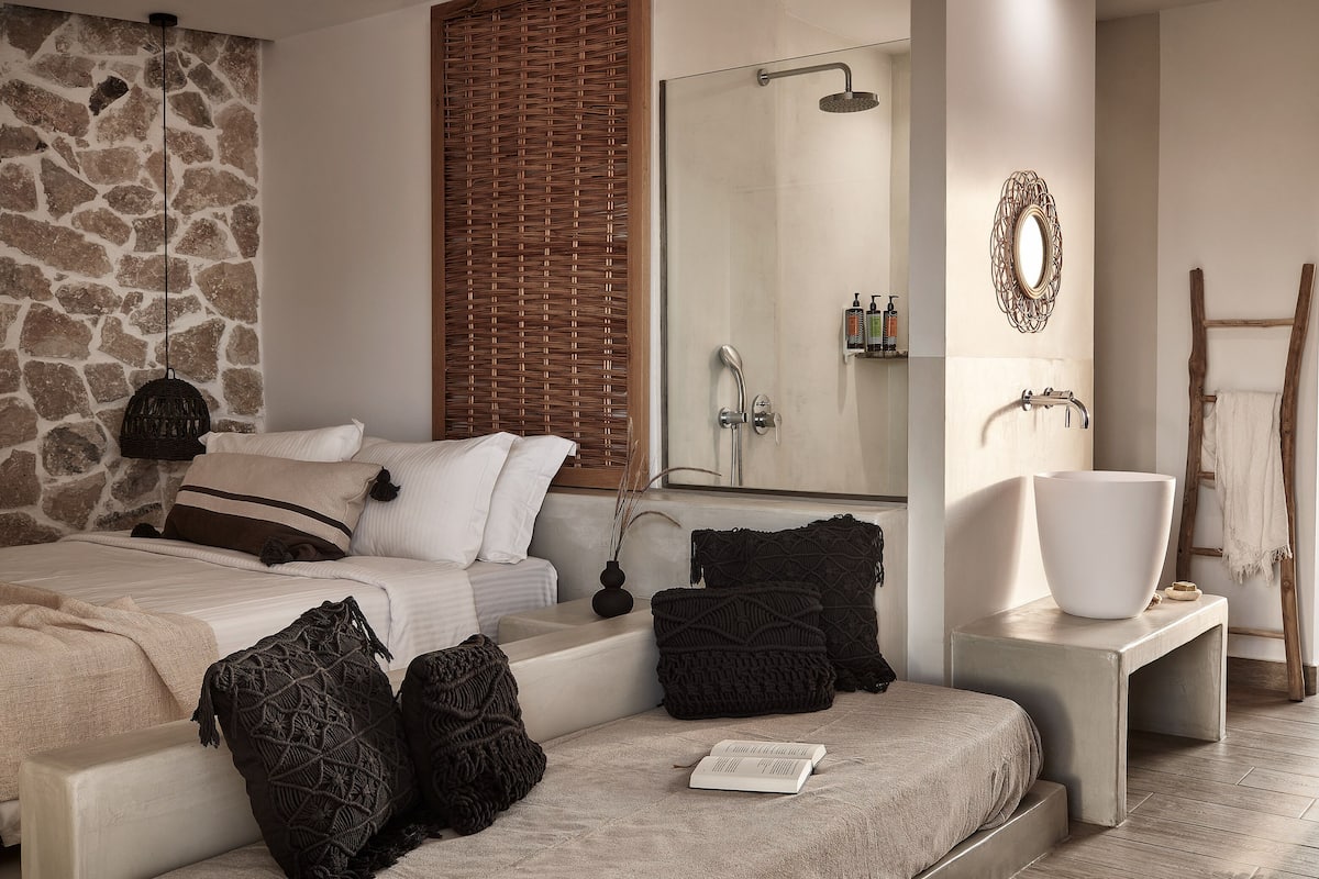 Un dormitorio con pared de piedra, una cama grande con muchas almohadas, una mesita de noche con un jarrón blanco y una escalera de madera apoyada en la pared.