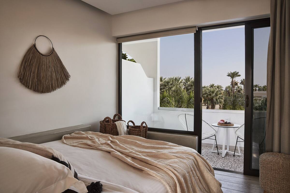 Un dormitorio con una cama grande, un adorno colgante en la pared, un balcón con mesa y sillas, y vistas a palmeras.