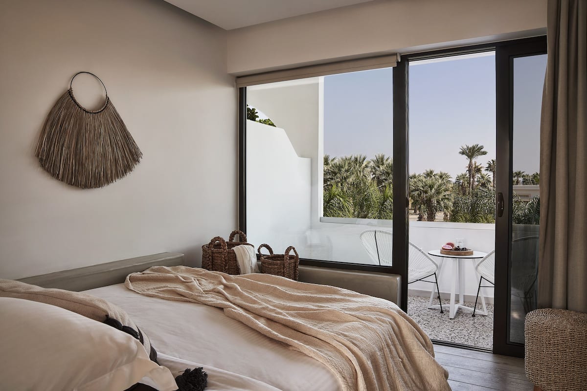 A bedroom with a large bed, a hanging wall decoration, a balcony with a table and chairs, and a view of palm trees.
