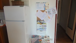 Una cocina con un refrigerador blanco, un tablón de anuncios con varios papeles y avisos, y una pequeña mesa con objetos encima.