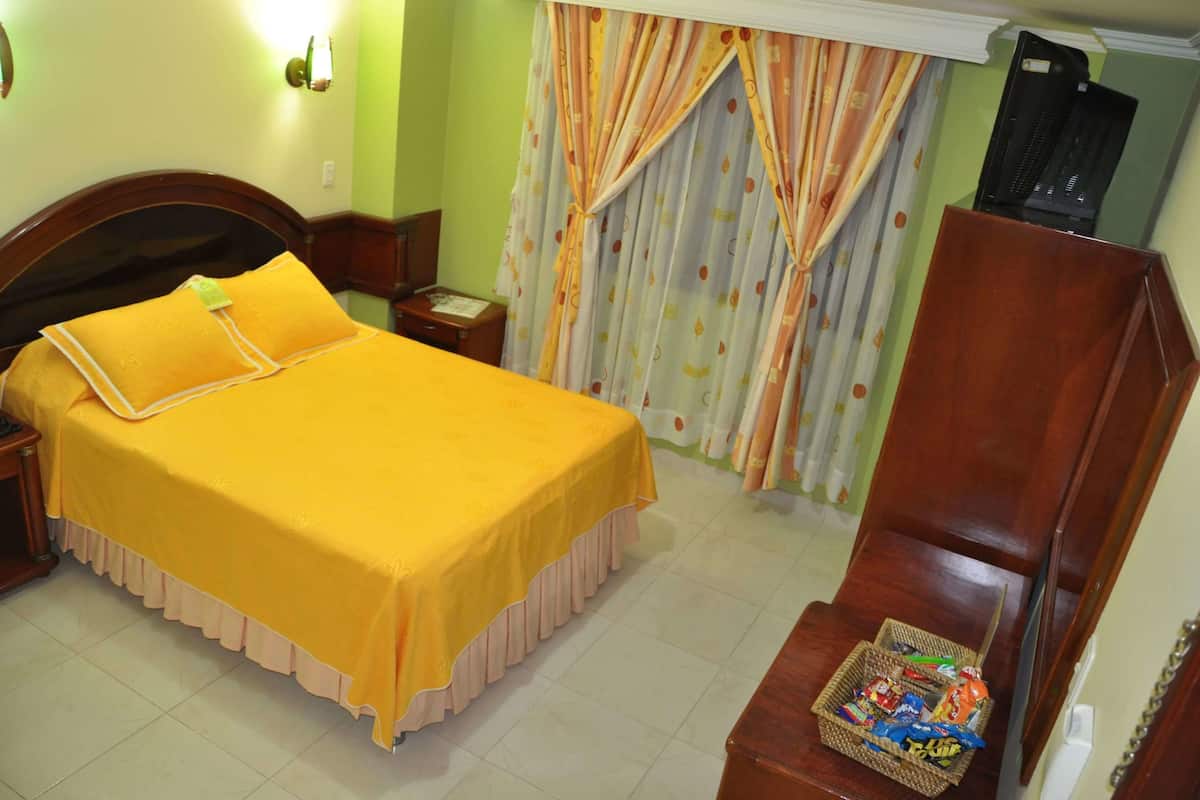Una cama bien hecha con un edredón amarillo brillante y almohadas, una mesita de noche de madera con una lámpara, una pequeña mesa con un teléfono y una cesta de mimbre con aperitivos.