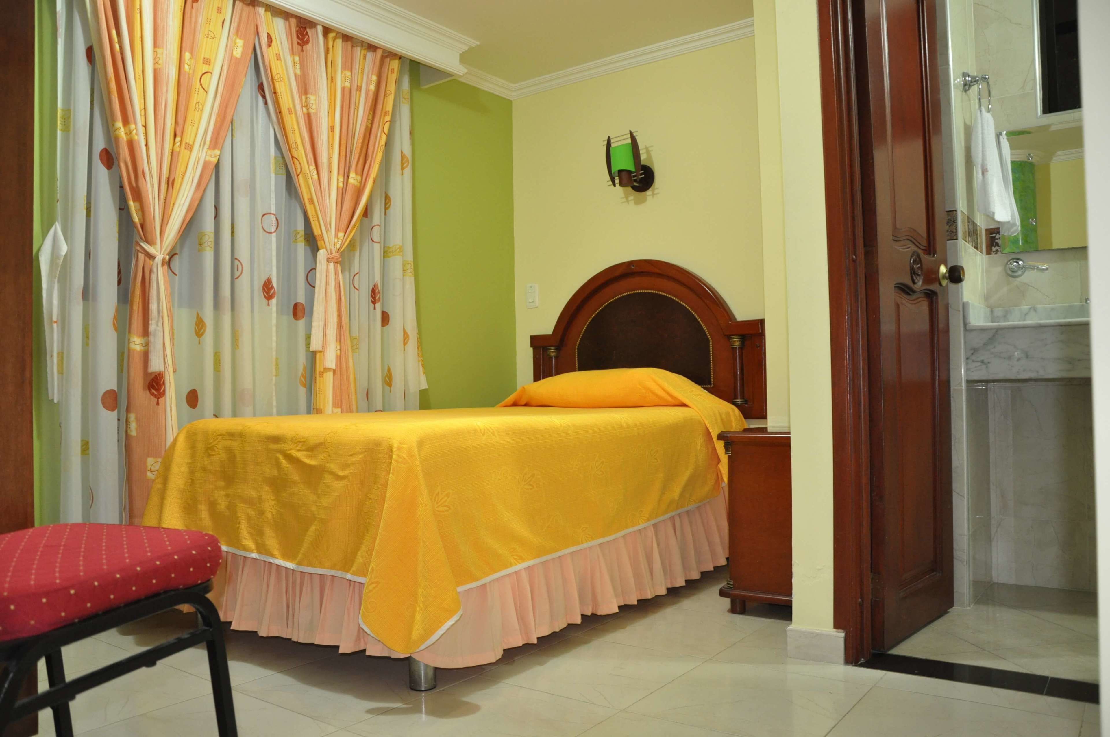 A room with a bed, a red chair, a wooden headboard, a bathroom with a mirror, and a door.