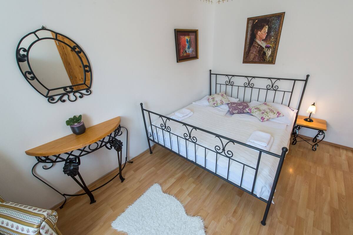 Quarto bem arrumado com estrutura de cama de metal, mesa de cabeceira de madeira com uma planta, tapete branco e fofo e uma pequena mesa de cabeceira com abajur.