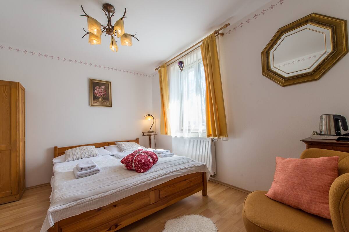 Quarto com cama de madeira, mesa de cabeceira, uma cadeira, um espelho e janela com cortinas.