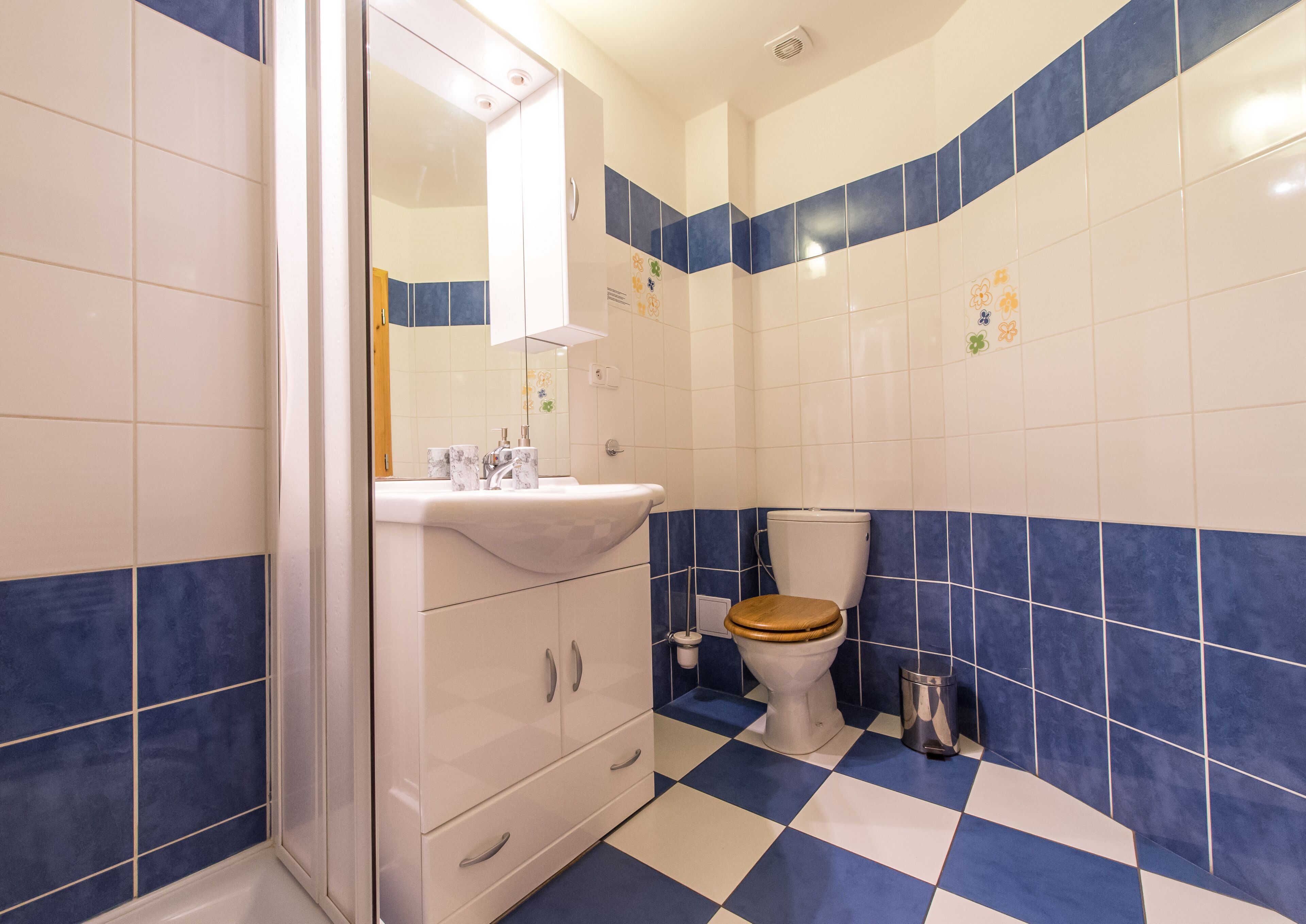 Un baño con un lavamanos blanco, un espejo encima, un inodoro con asiento de madera y azulejos blancos y azules en las paredes y el piso.