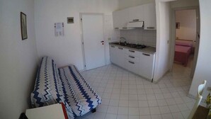 Eine kleine Küche mit weißen Schränken, einem Gasherd und einem Spülbecken. Im angrenzenden Raum befindet sich ein Bett mit einem gemusterten Überwurf.