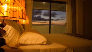 A hotel room with a bed, a nightstand, and a view of the sunset through a large window. - The Streamliner Hotel Apartment (Toamasina)