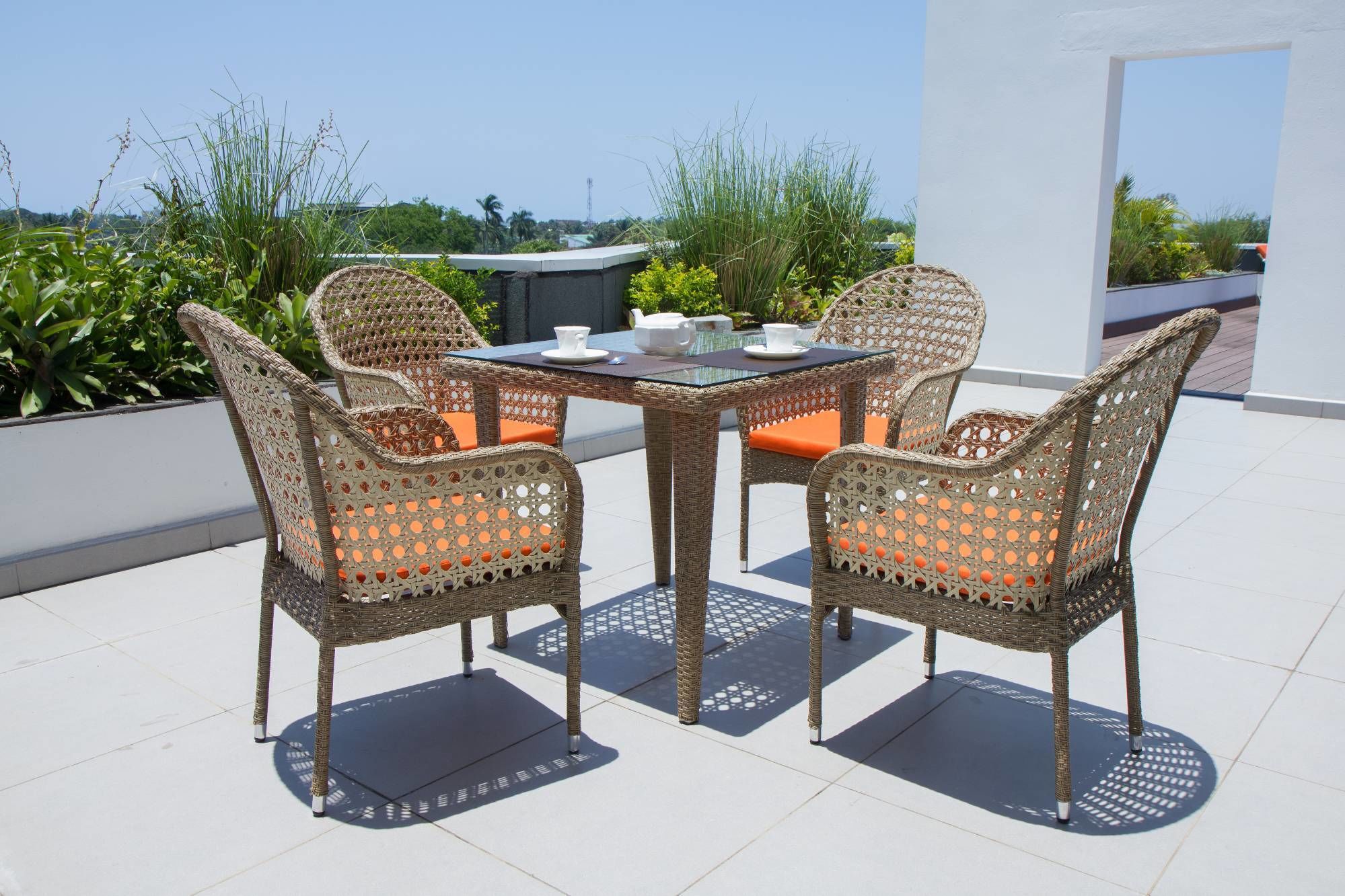 A wicker outdoor dining set with orange cushions, a glass table, and white teacups on a rooftop terrace with a view of greenery and buildings.