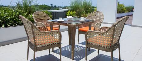 A wicker outdoor dining set with orange cushions, a glass table, and white teacups on a rooftop terrace with a view of greenery and buildings.