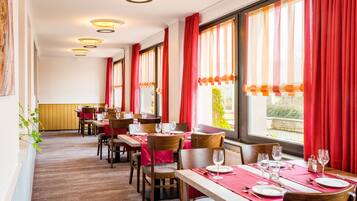 Een restaurant met meerdere eettafels, rode gordijnen en uitzicht op groen.