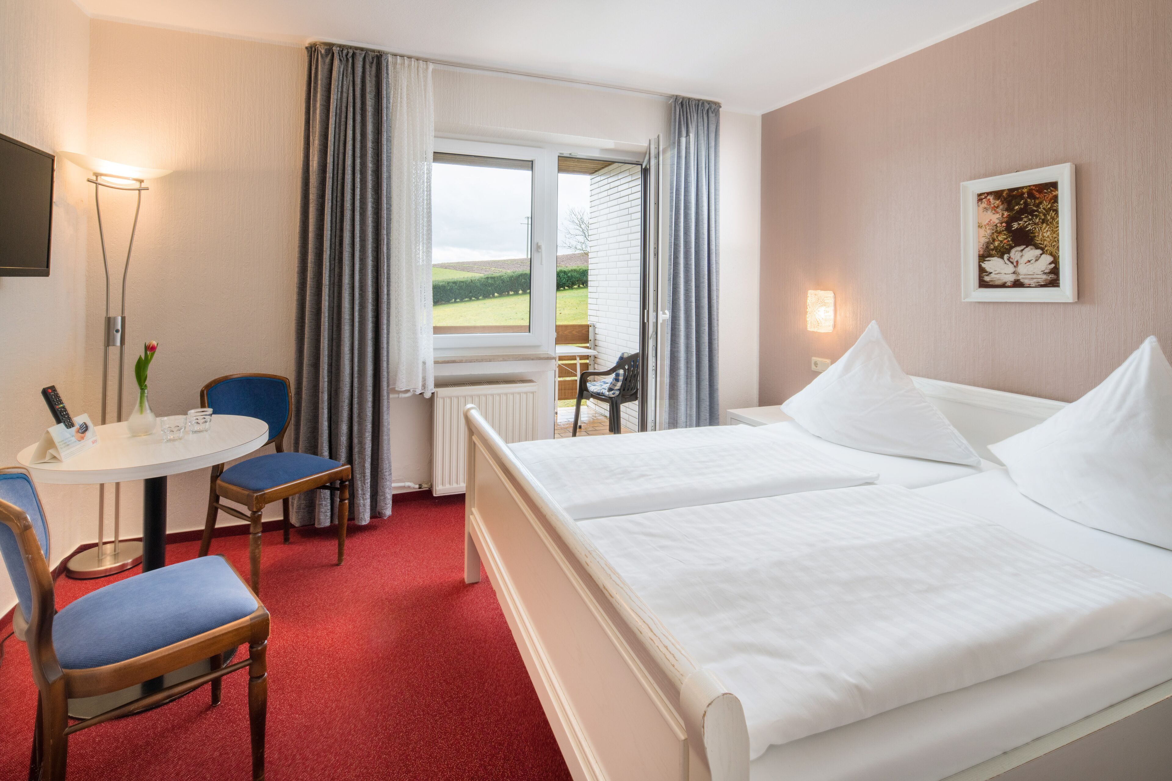 A hotel room with two beds, a small table with a chair, a radiator, and a window with curtains.