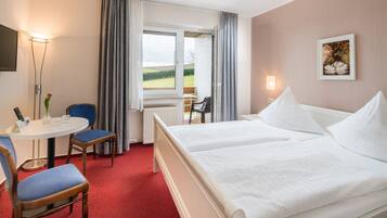A hotel room with two beds, a small table with a chair, a radiator, and a window with curtains.
