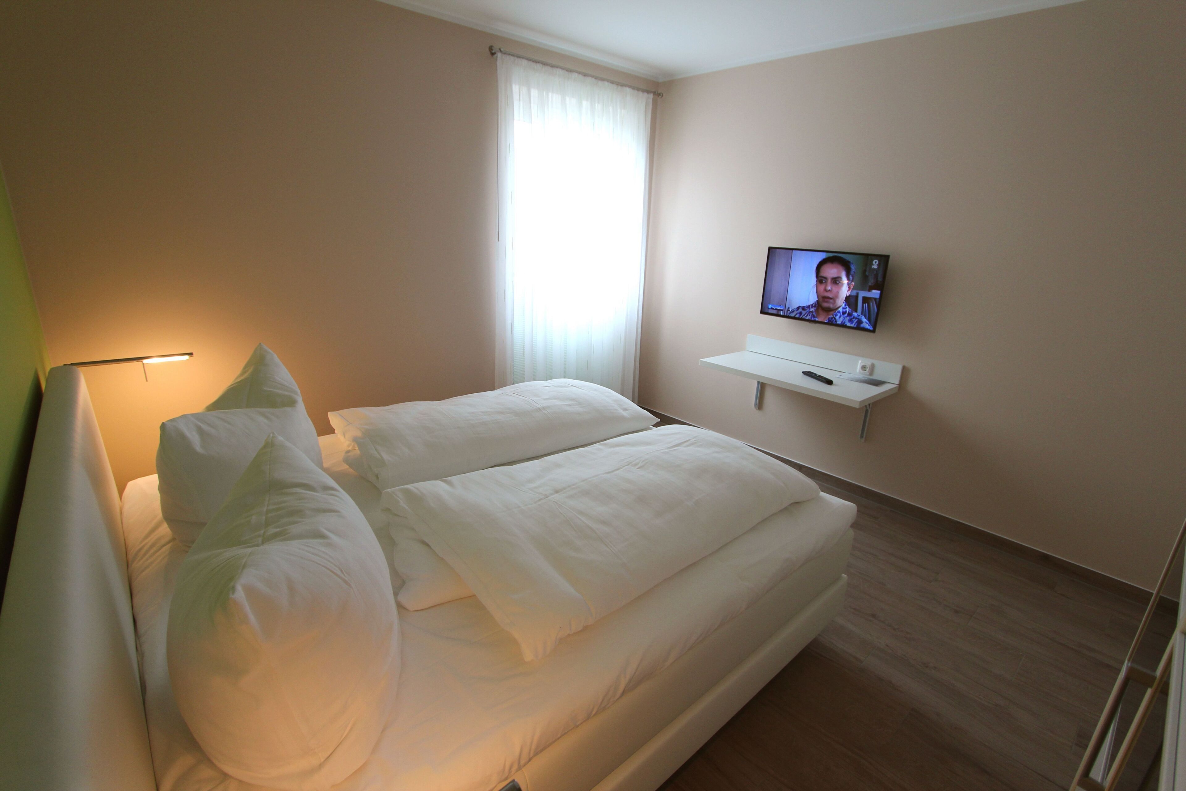 Hotellihuone, jossa on suuri sänky, seinälle asennettu taulutelevisio ja ikkuna, jossa on ohuet verhot.