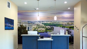 A reception desk with a mural of a cityscape and river, two pendant lights, and a small plant on the counter. - Americas Best Value Inn & Suites Prairieville (Prairieville)