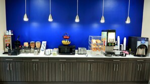 A hotel reception area with a blue wall, a coffee machine, a microwave, a toaster, and various food items on the counter. - Americas Best Value Inn & Suites Prairieville (Prairieville)